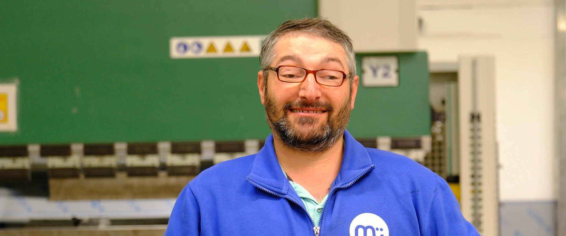 Mann mit Brille und Bart in blauer Jacke mit Logo hält Akkuschrauber in der Metallabteilung einer Werkstatt